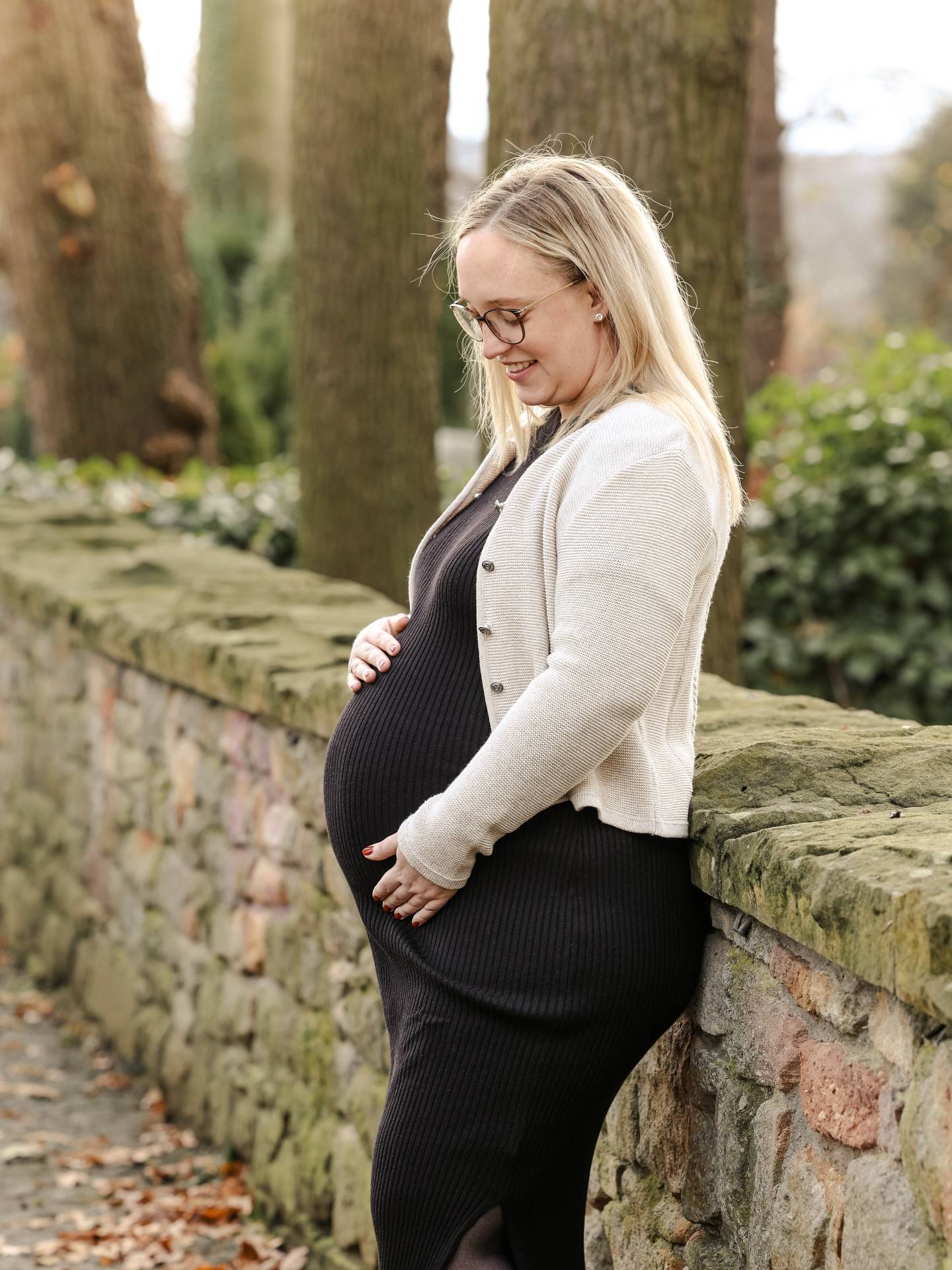 Schwangere Frau in dunklem Kleid und heller Strickjacke lehnt an einer moosigen Bruchsteinmauer und schaut lächelnd auf ihren Babybauch.