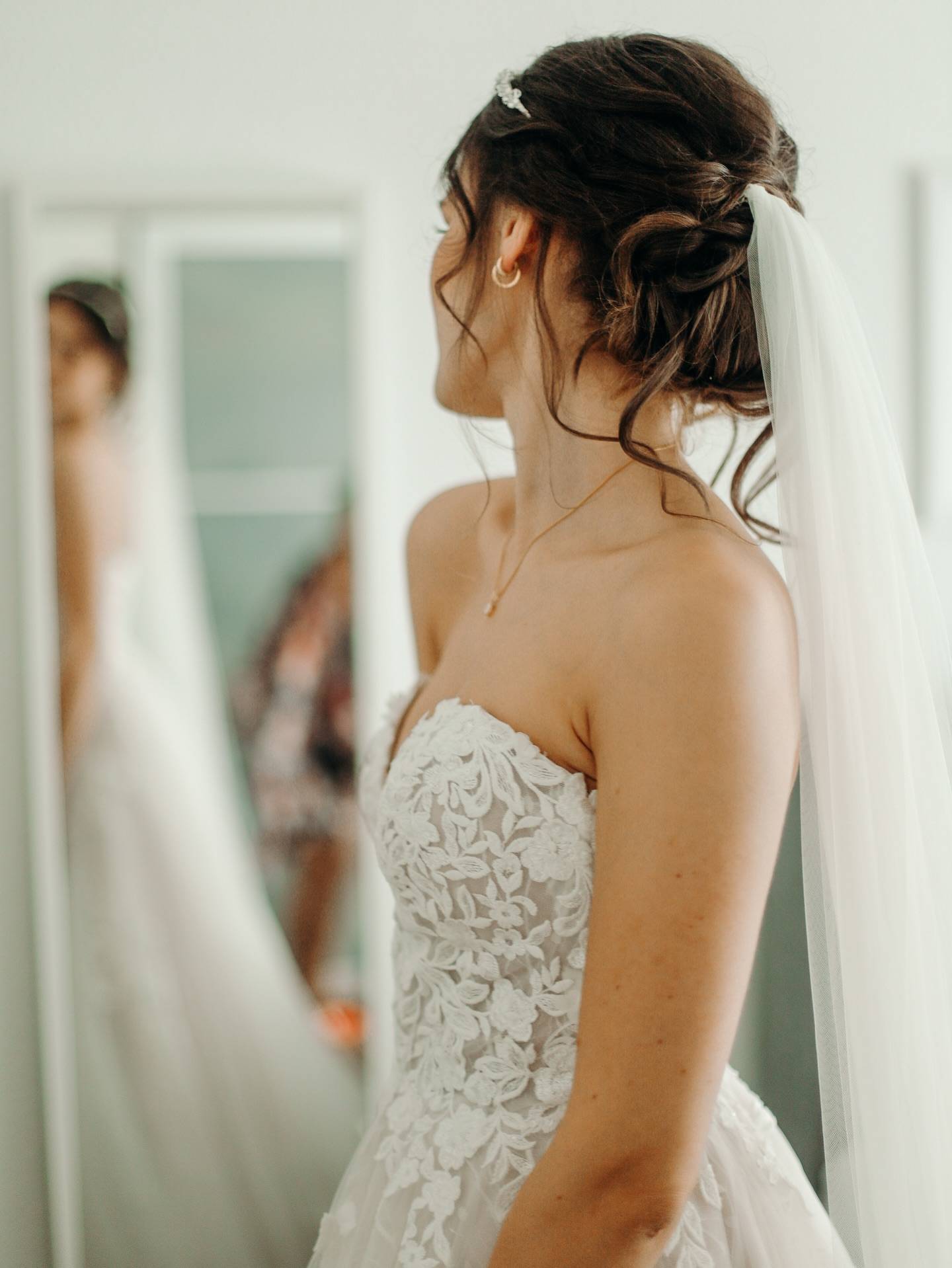 Braut im trägerlosen Spitzenkleid mit Schleier und hochgestecktem Haar schaut zur Seite, links spiegelt sie sich.