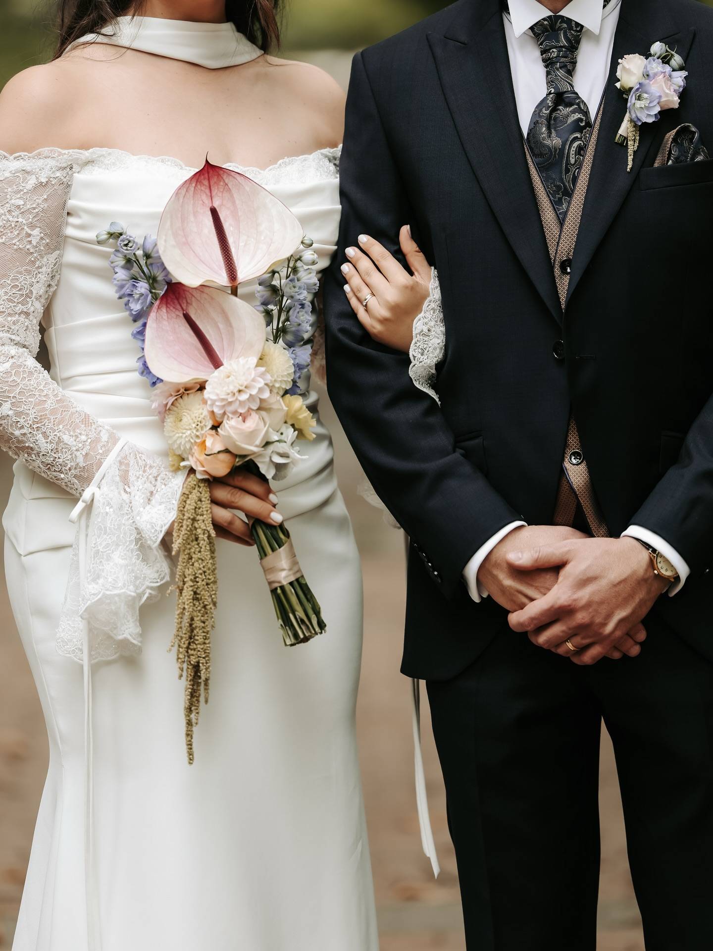 Braut im Spitzenkleid mit Brautstrauß aus Anthurien und Trockengräsern hakt sich beim Bräutigam im schwarzen Anzug ein.