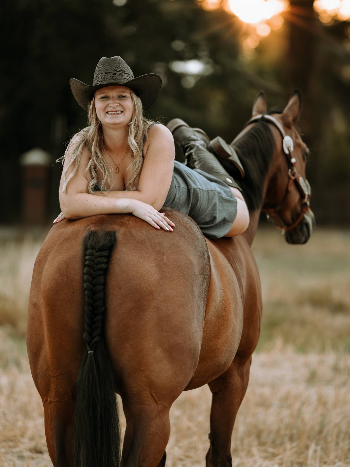 Lachende Frau mit Cowboyhut liegt bäuchlings quer über dem Rücken eines braunen Pferdes mit geflochtenem Schweif, dahinter die Abendsonne.