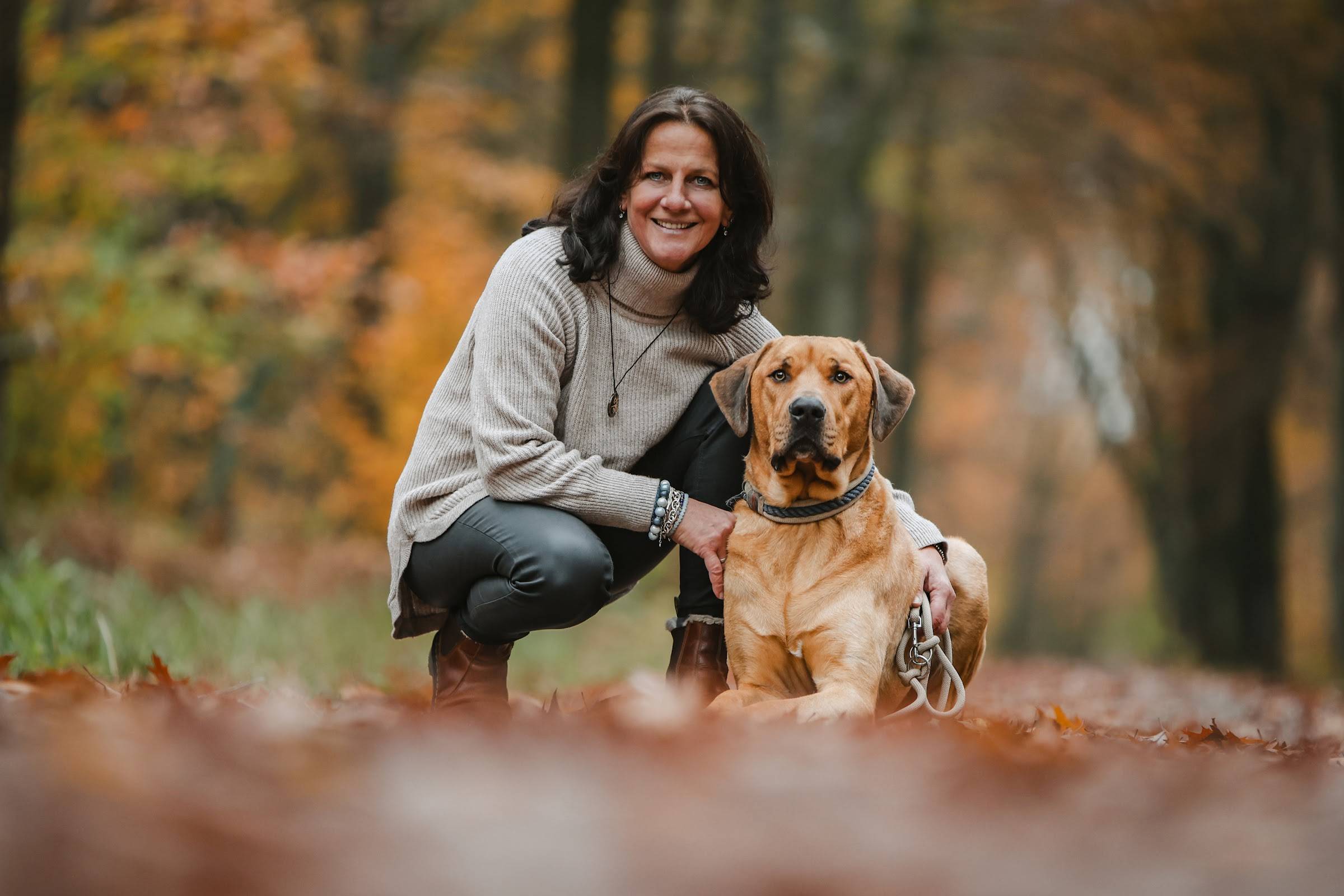 Frau im hellen Strickpullover hockt lachend neben ihrem großen hellbraunen Hund im Herbstlaub eines Waldes.