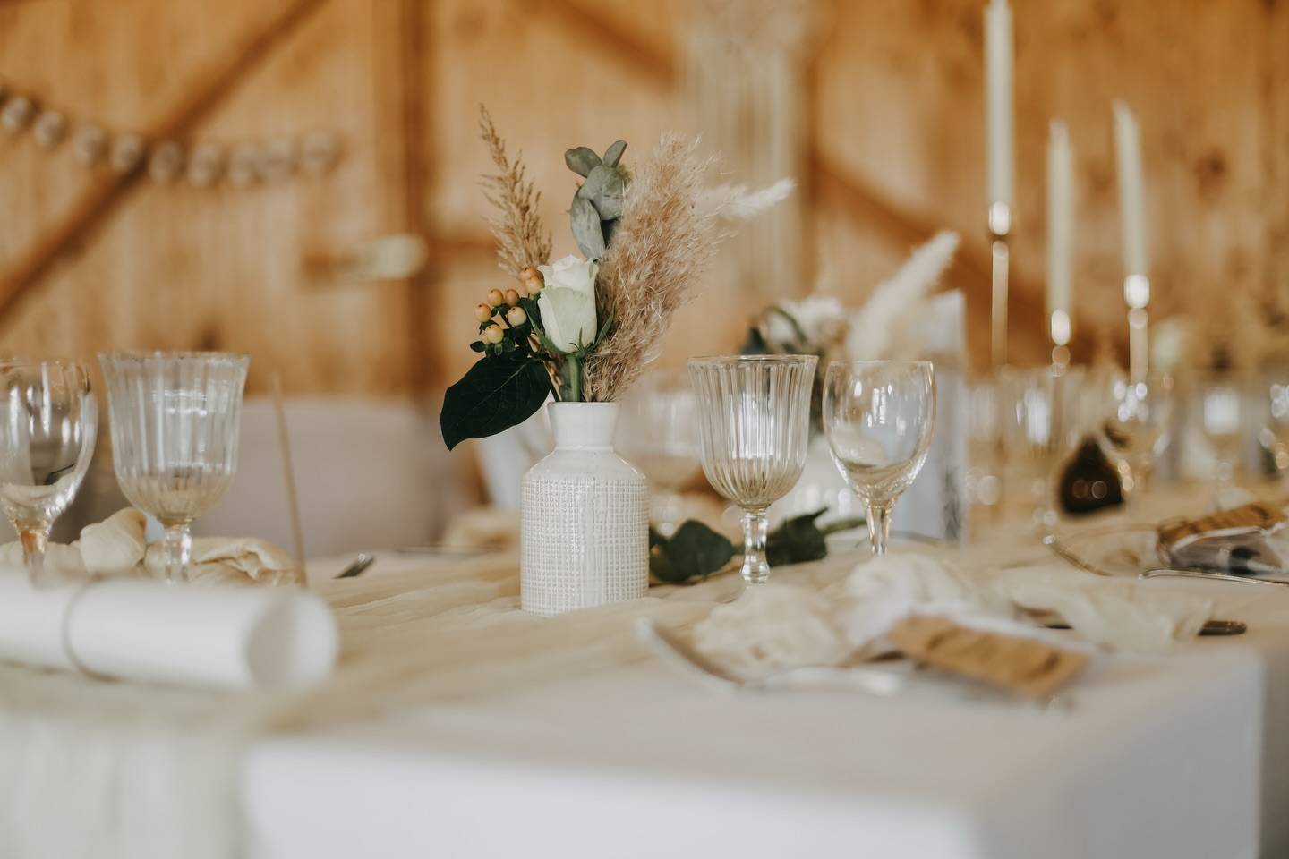 Festlich gedeckte Hochzeitstafel mit weißen Tischdecken, Gläsern, Kerzen und Trockenblumen vor einer Holzwand.
