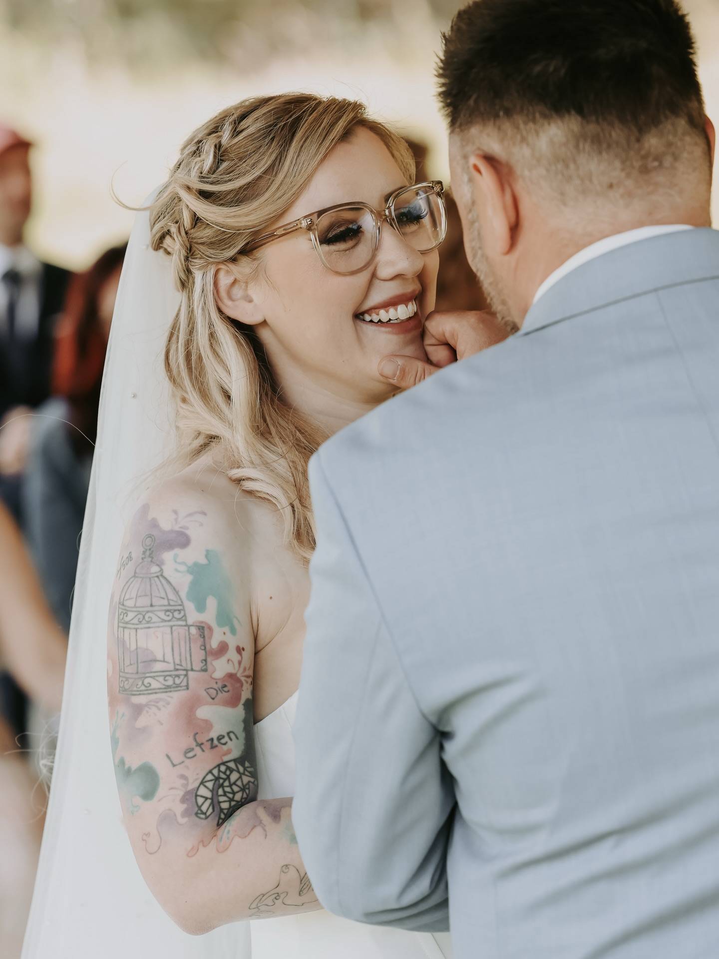 Braut mit Brille und Blumen im Haar lacht während der Trauung, der Bräutigam im hellblauen Anzug steht ihr zugewandt.