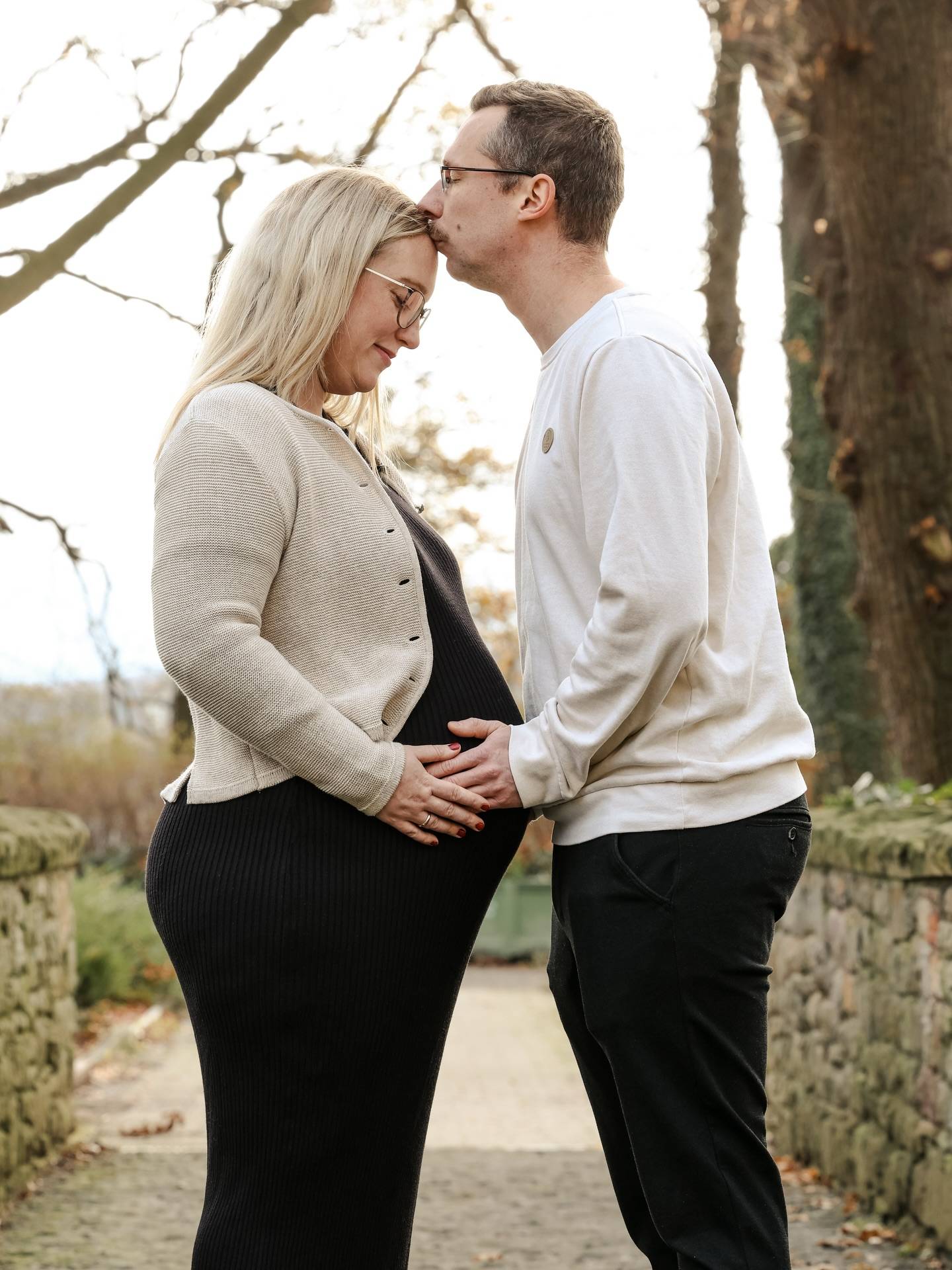 Werdende Eltern stehen im Park, er küsst ihre Stirn, beide halten die Hände auf ihrem Babybauch.